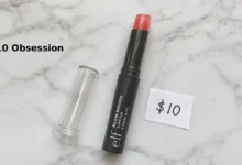 e.l.f. Glow Reviver Slipstick in shade Coral Kiss. The slim twist-up tube with clear cap is shown on a clean white surface. The product is partially twisted up to show the squishy gel texture. A small handwritten tag says "$10". Soft natural lighting.