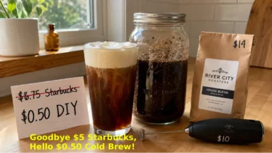 A glass of homemade cold brew with vanilla sweet cream cold foam on top. Next to the glass, a Mason jar with coffee grounds and water steeping, a bag of coffee beans, and a handwritten card showing "$0.50 vs $6.75".