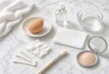 Close-up of a makeup sponge with a spritz of water, a clean tissue, and a damp cotton cloth next to a compact mirror – DIY free household fixes for cakey foundation.