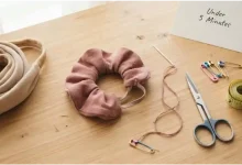 A stretched-out fabric scrunchie next to a needle, matching thread, and a pair of scissors on a wooden table – quick no-sew and sewing hacks to fix a stretched hair tie in 5 minutes.