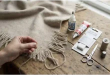 A person's hands hand-sewing the frayed fringe of a beige herringbone scarf on a rustic wooden table, next to a bottle of Fray Check, fabric glue, iron-on hem tape, scissors, a spool of thread, and a thimble.