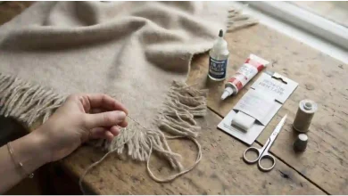 A person's hands hand-sewing the frayed fringe of a beige herringbone scarf on a rustic wooden table, next to a bottle of Fray Check, fabric glue, iron-on hem tape, scissors, a spool of thread, and a thimble.